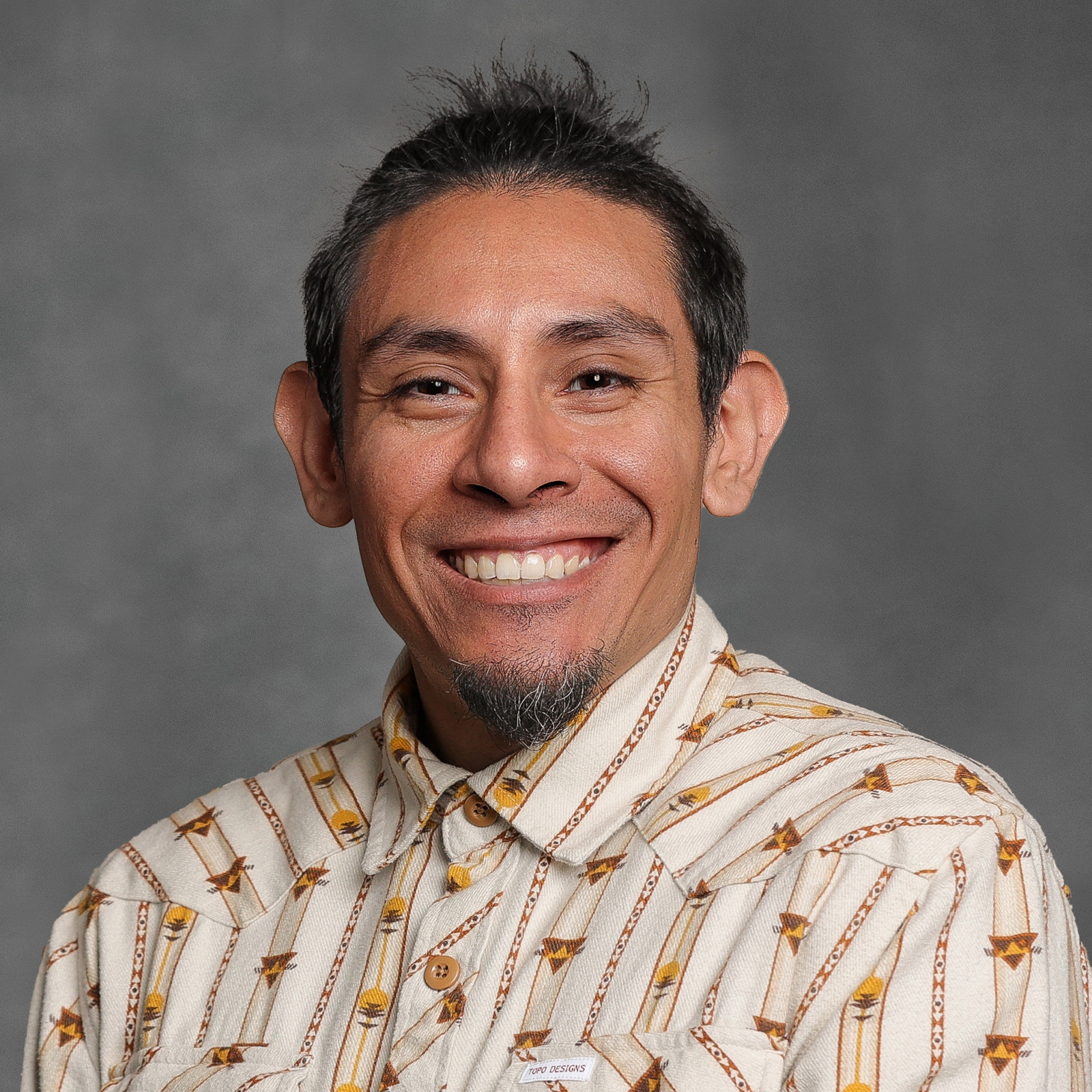 Man with short, dark hair and chin patch wearing tan shirt with brown and gold print