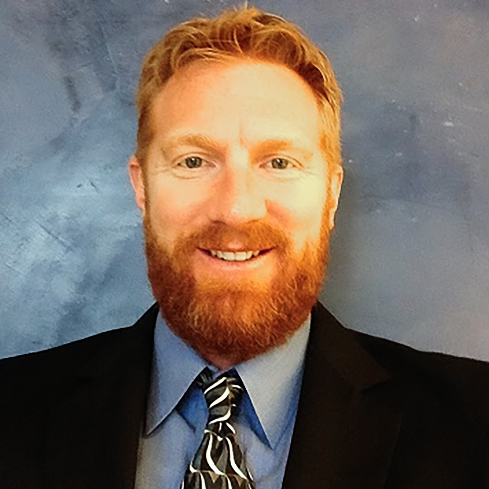 Man with red hair and beard in dark suit, blue shirt and black print tie 