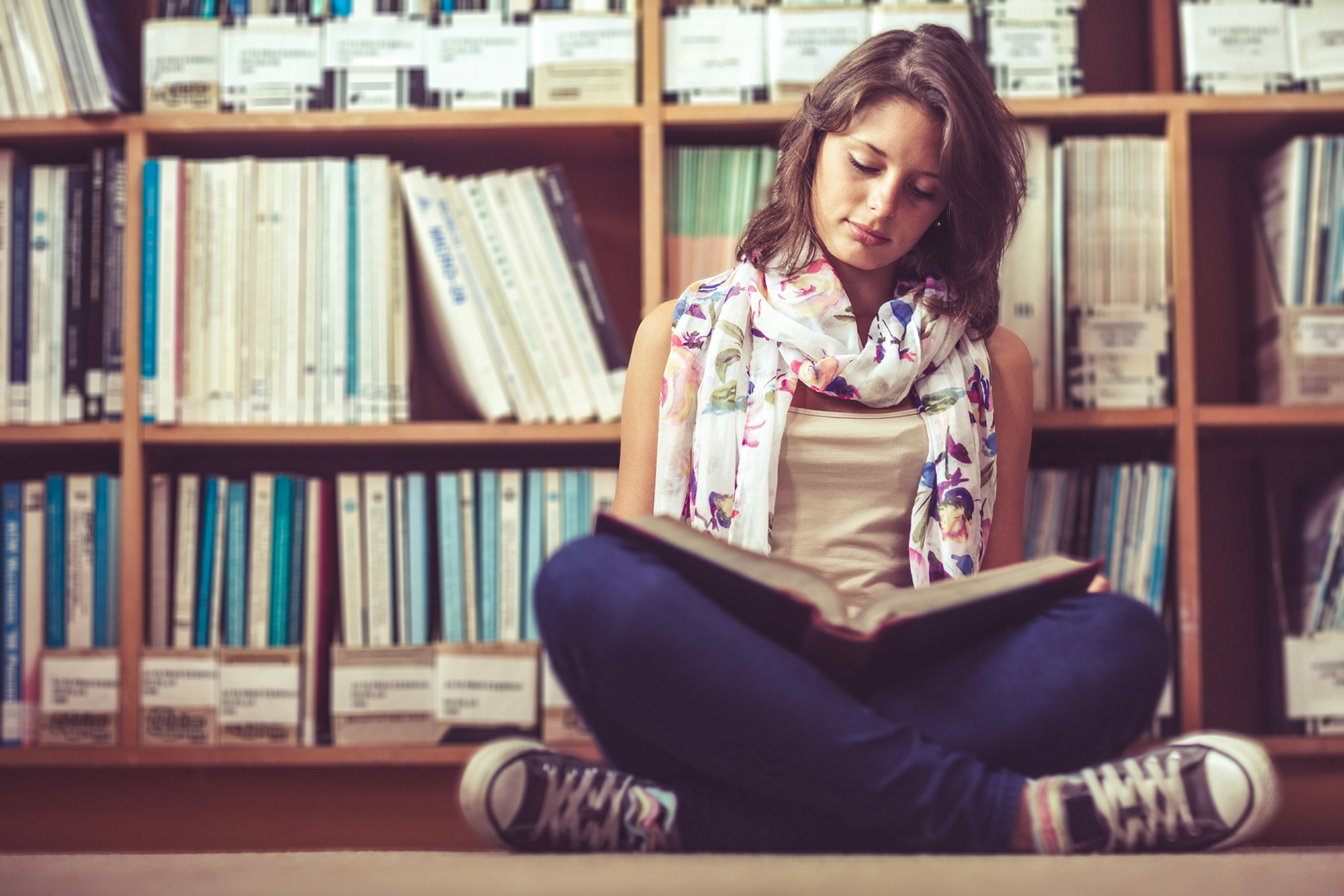 Woman sitting cross legged on the floor of the library reading a book.