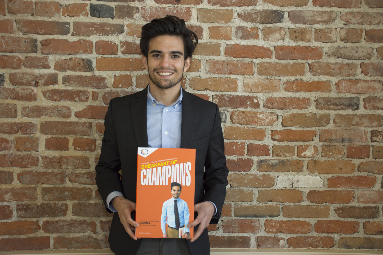 CCD Associate of Arts – Communication Student Saul Mejia, CCD Foundation Open Door Scholarship young man holding a box with his picture on it, in front of a brick background