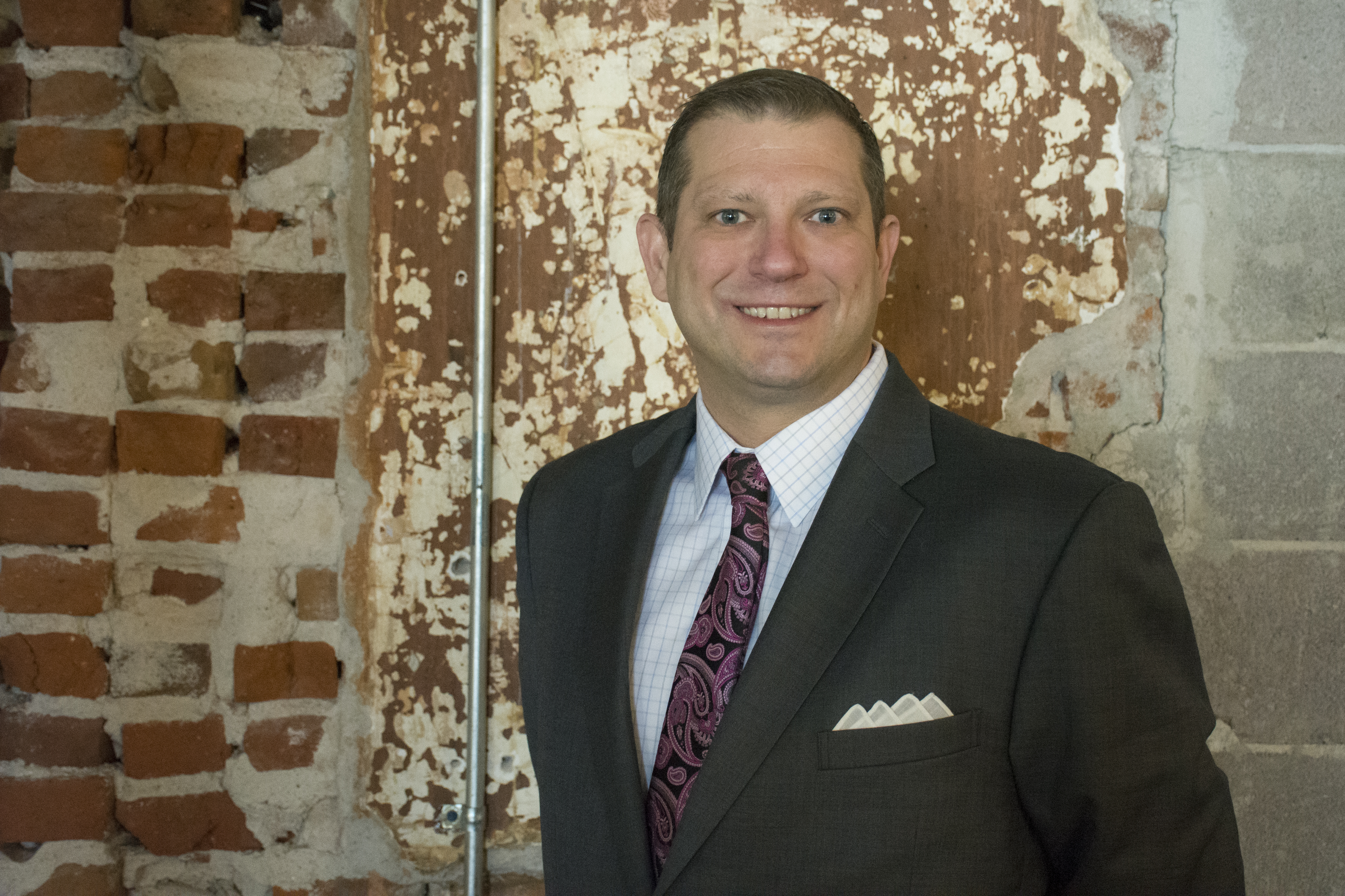 white man in a suit standing in front of a brick wall smiling