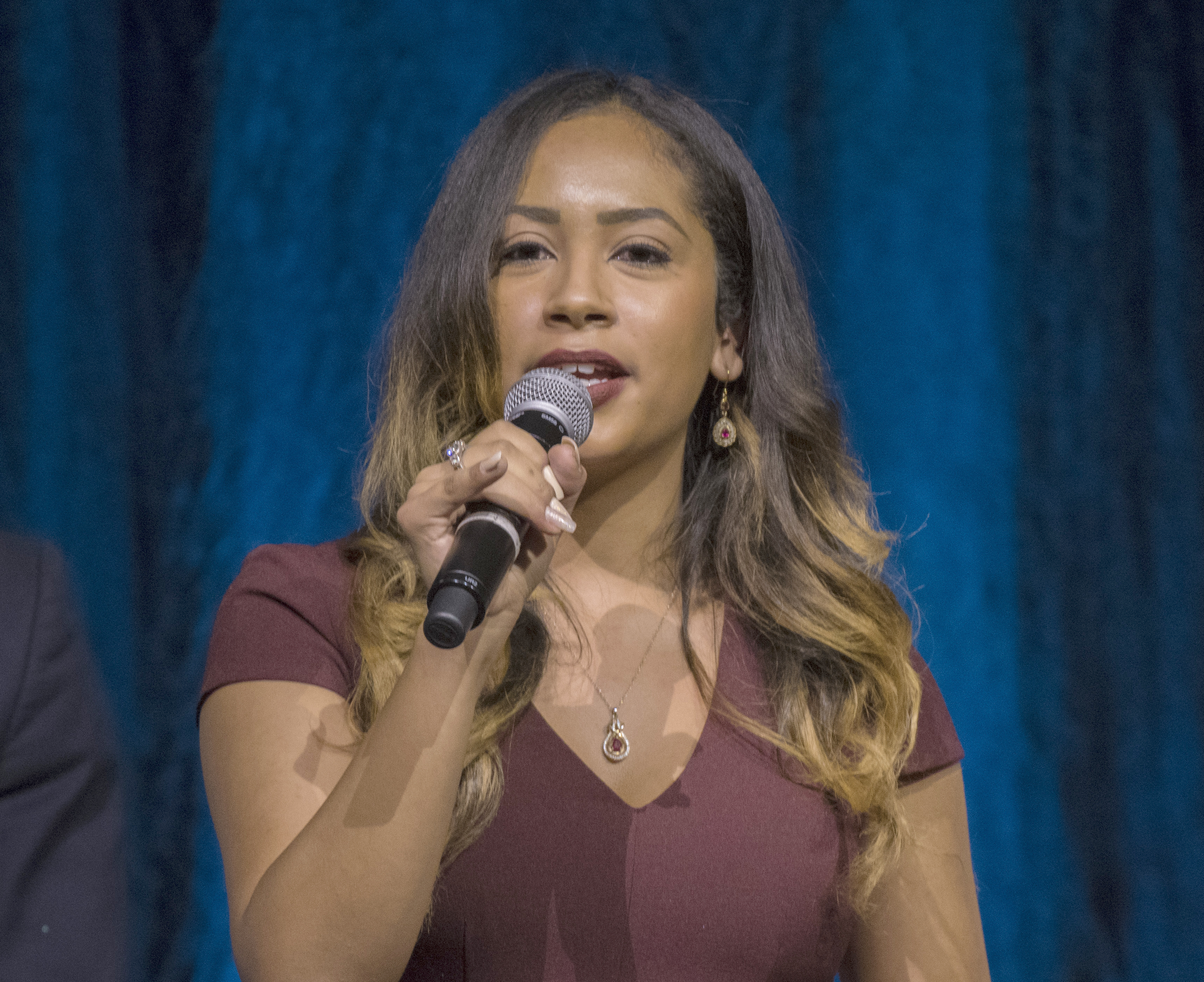 Hispanic woman in a maroon dress speaking into a microphone