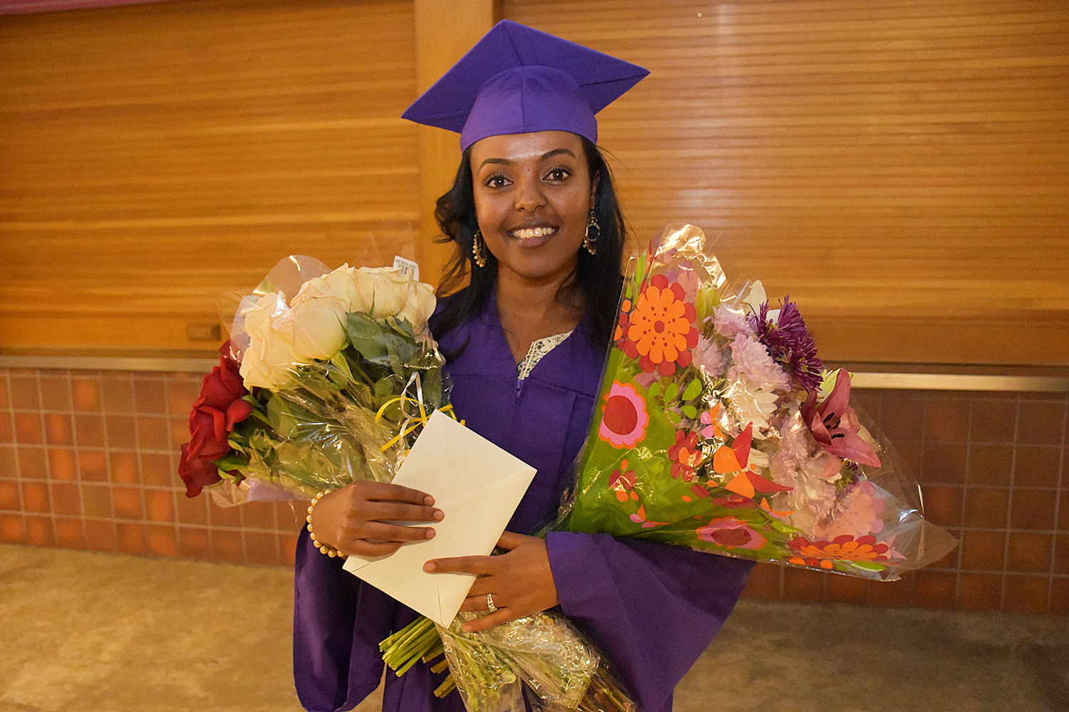 smiling woman in purple cap and gown holds celebratory flowers
