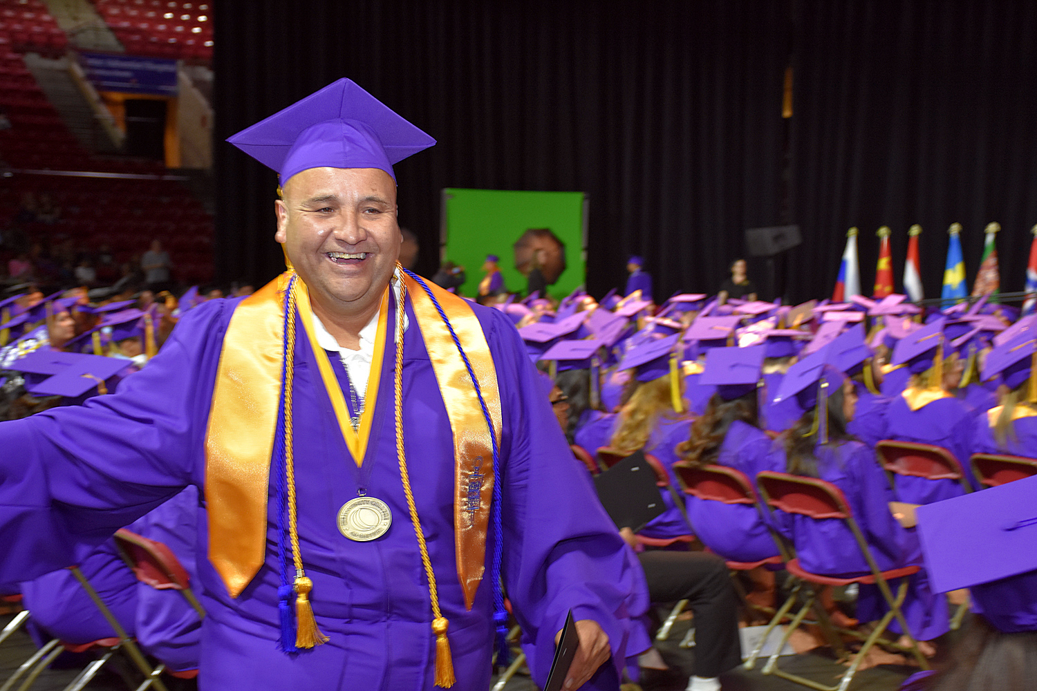 smiling man in purple cap and gown and gold honor society sash receives his diploma
