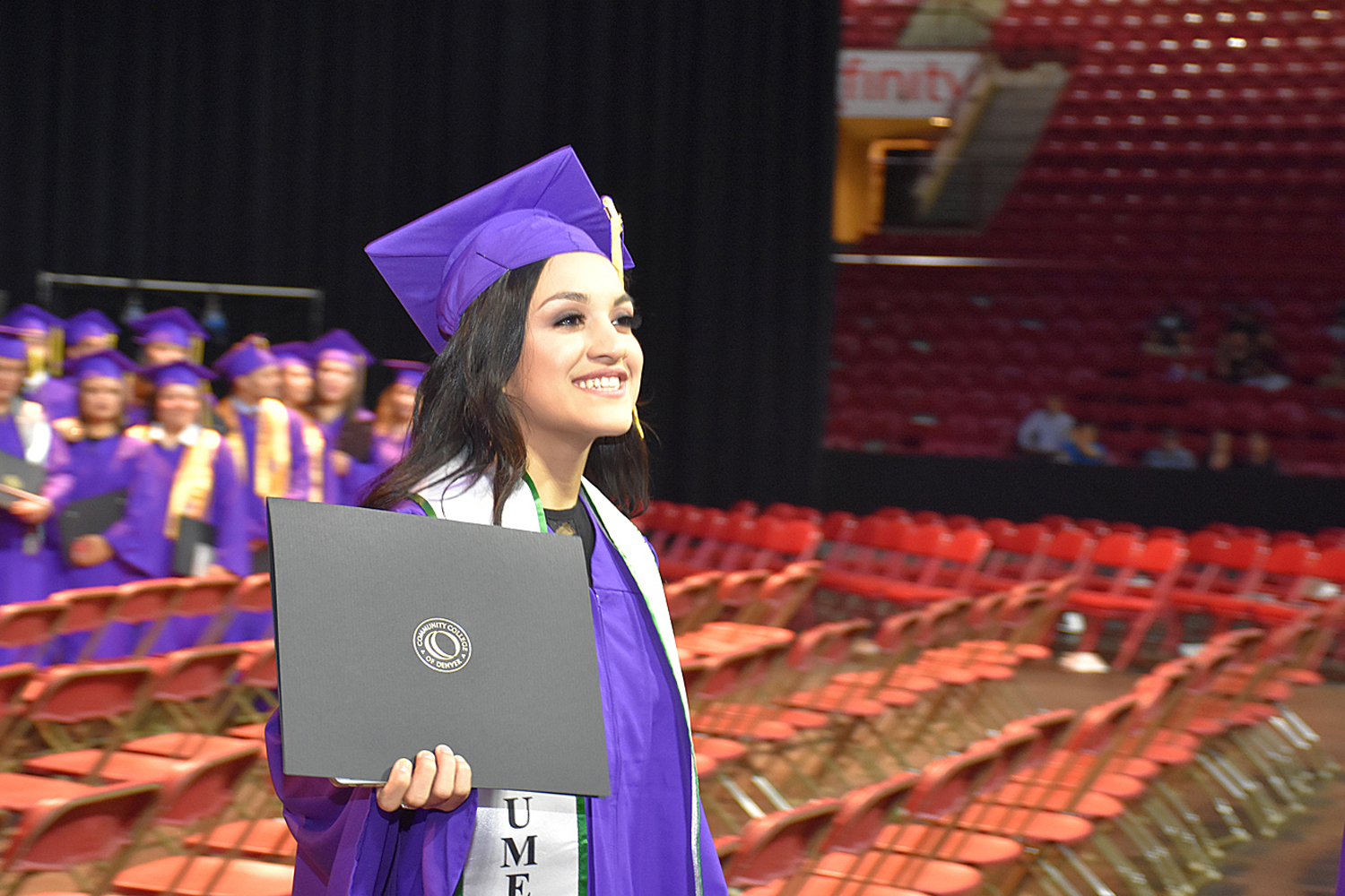 woman in purple cap and gown holds up a black envelope with CCD logo and diploma