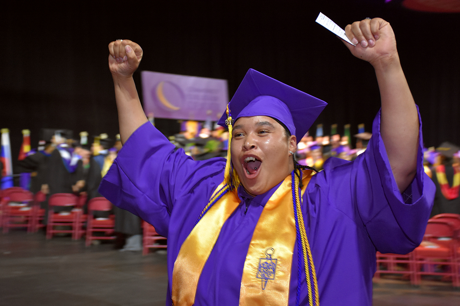 student in purple cap and gown and gold honor society sash holds is hands in the air in celebration