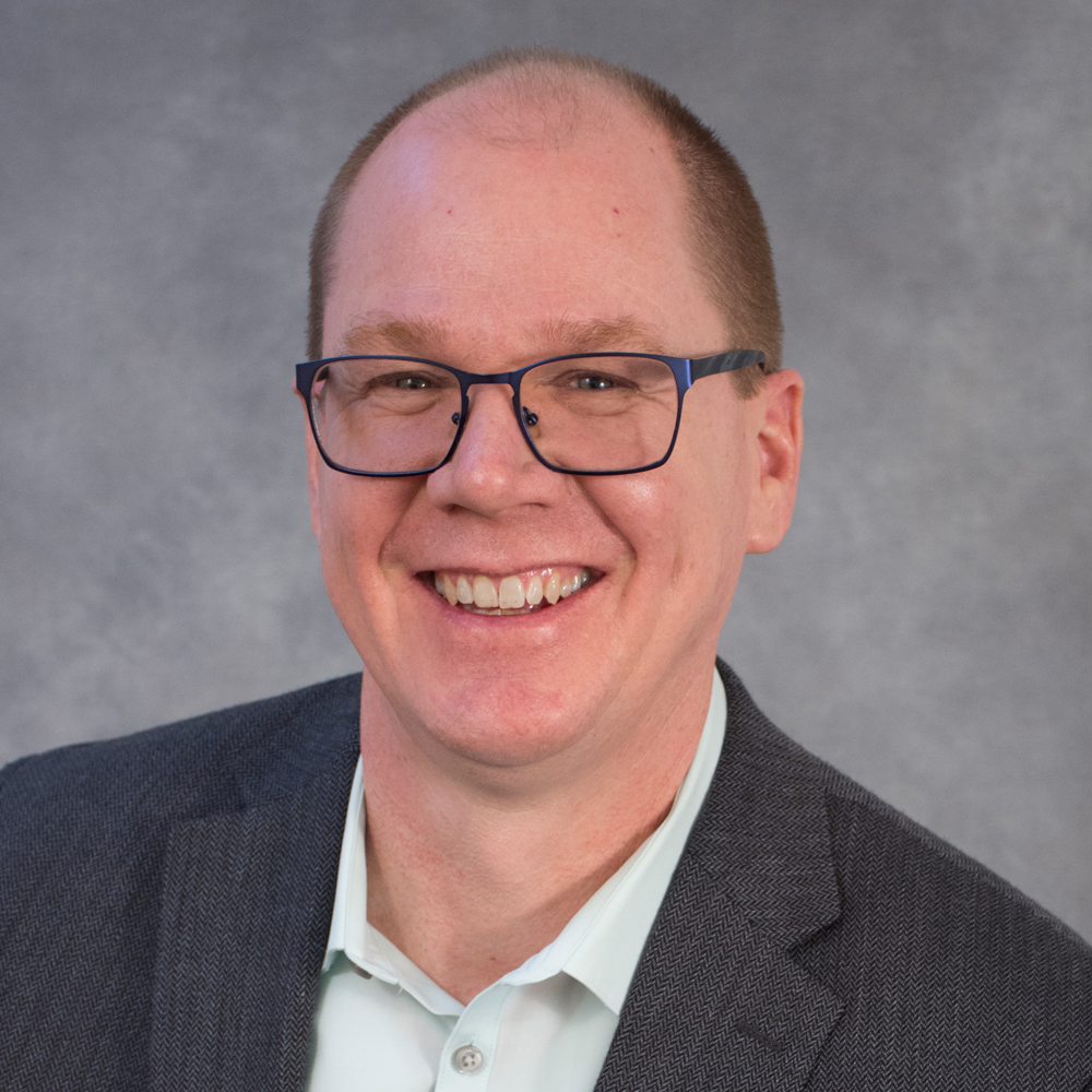 Smiling man with cropped short brown hair wearing glasses and gray jacket