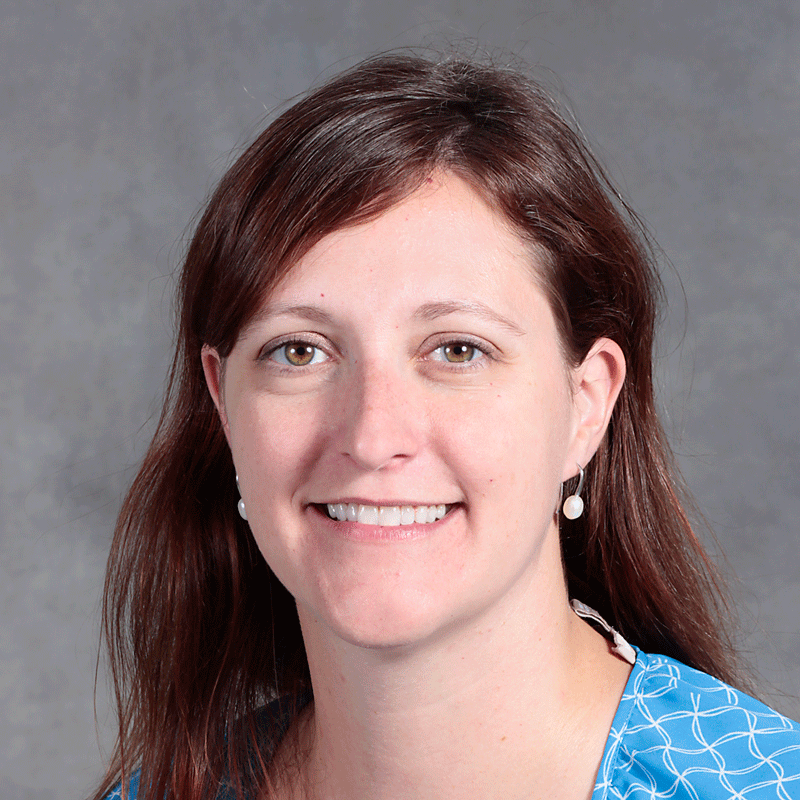 smiling woman with shoulder length brown hair wearing blue top and pearl earrings