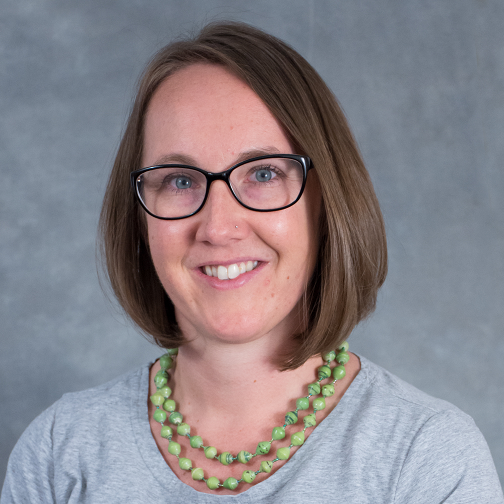 Smiling person with chin length brown hair wearing glasses, a gray top and green beads