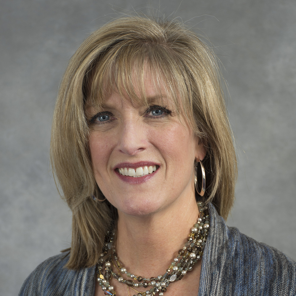 Smiling woman with medium length blond hair wearing grey top, beads and hoop earrings