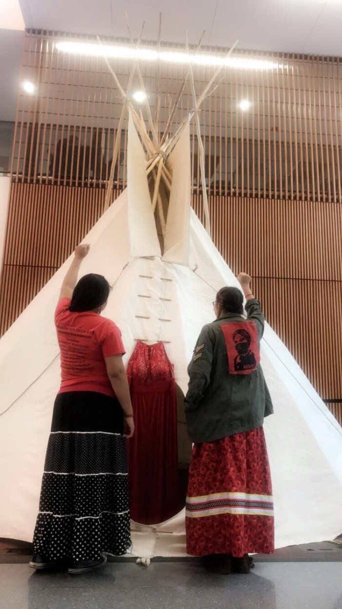 Two Females with fists raised in front of a tipi