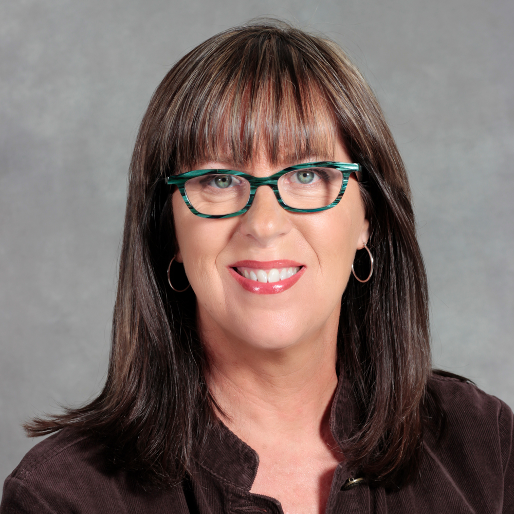 smiling woman with mid length dark brown hair, glasses and brown jacket