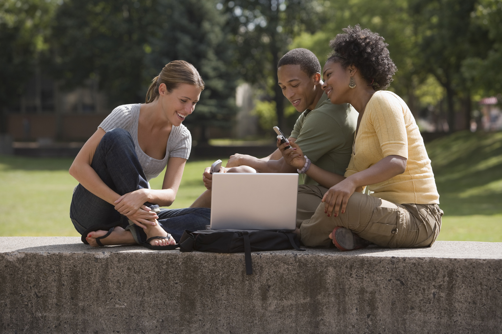 Two female students and one male student sitting outdoors looking at a laptop and phone smiling.