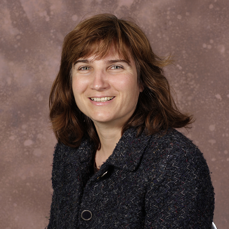 Smiling woman with medium length brown hair and bangs, wearing a dark gray jacket