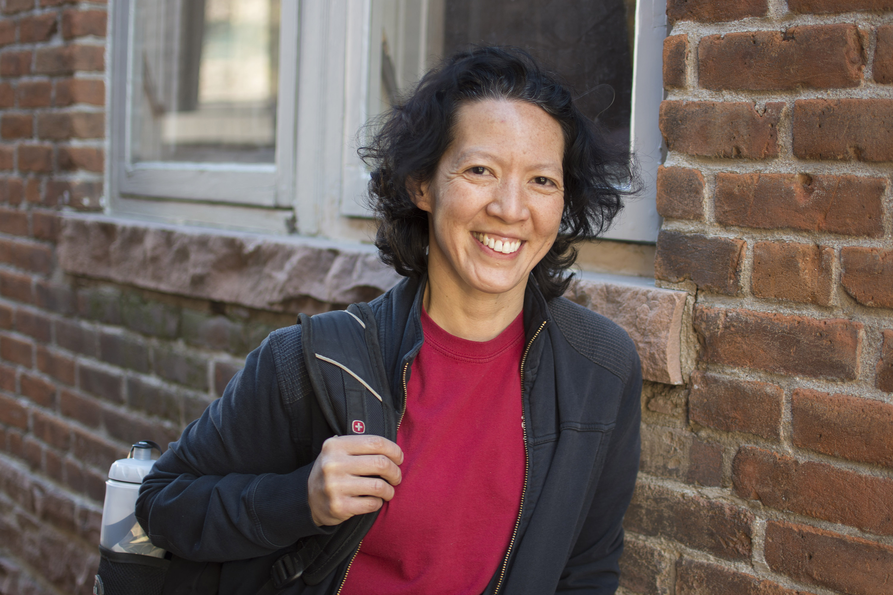 woman in a red shirt with a backpack over her shoulder smiling next to a brick wall