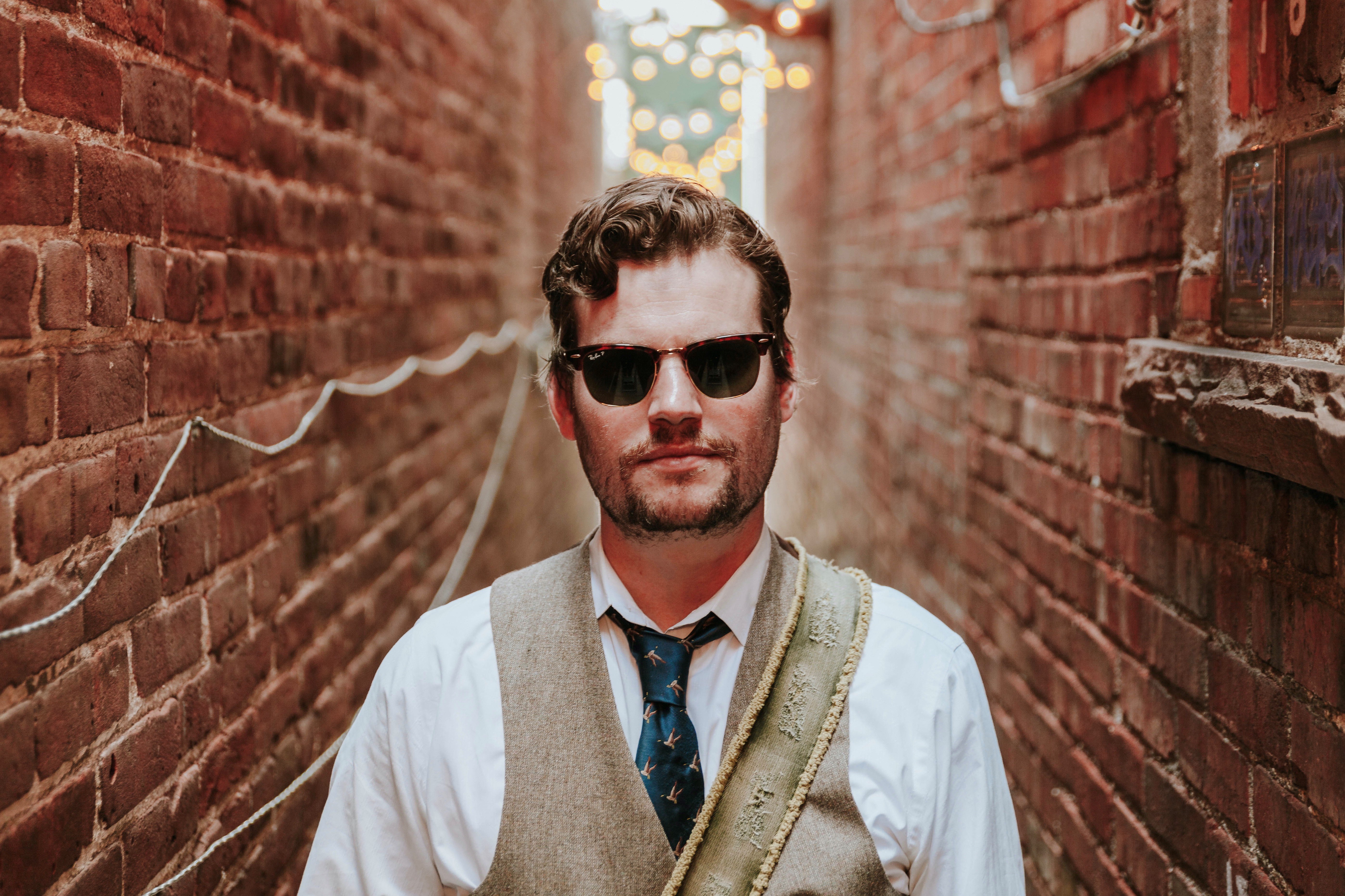 white man in sunglasses wearing a white fhirt and tan best standing in between two brick walls