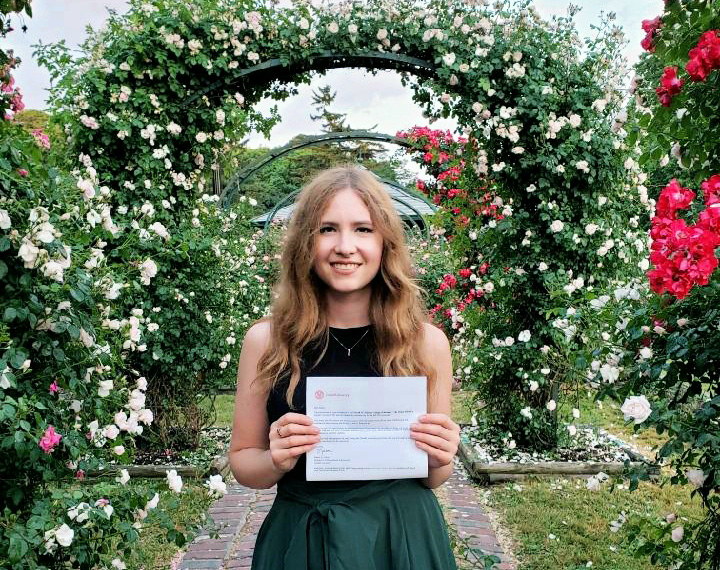 young white woman in a green dress holding a certificate in a garden surrounded by flowers