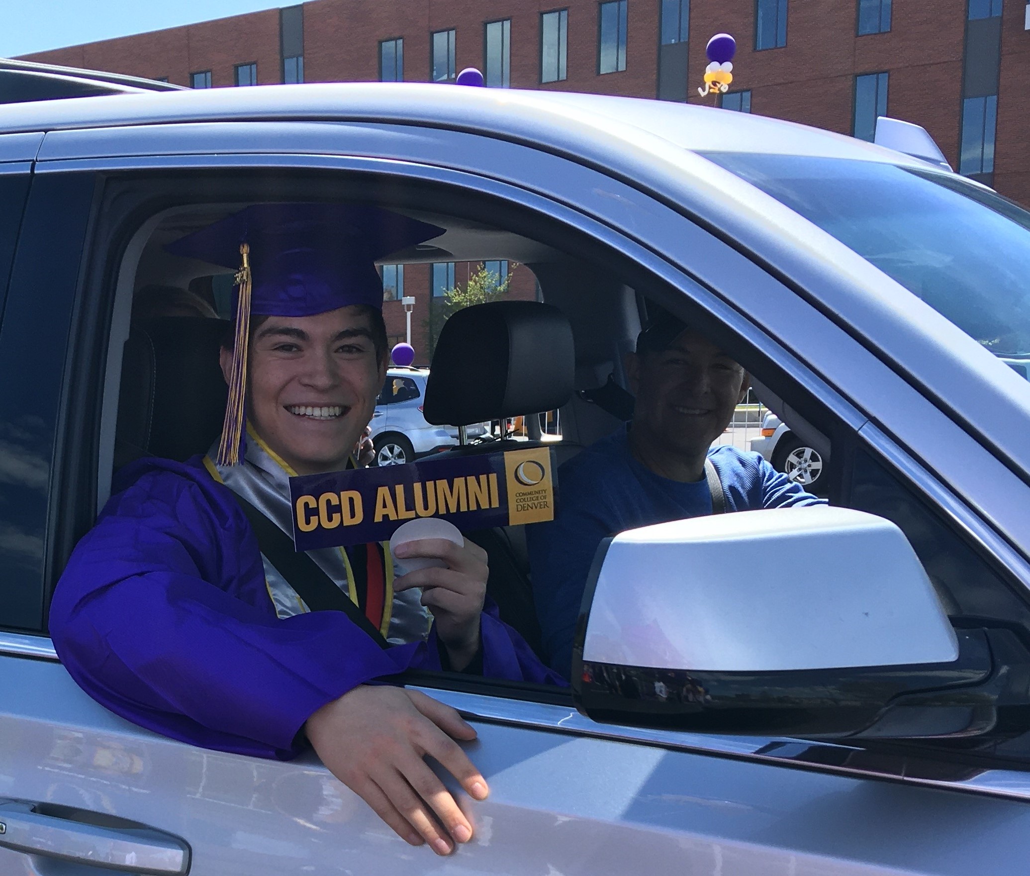 CCD graduate, Benjamin Rodriguez, with cap, gown and CCD Alumni bumper sticker, sitting in car at graduation