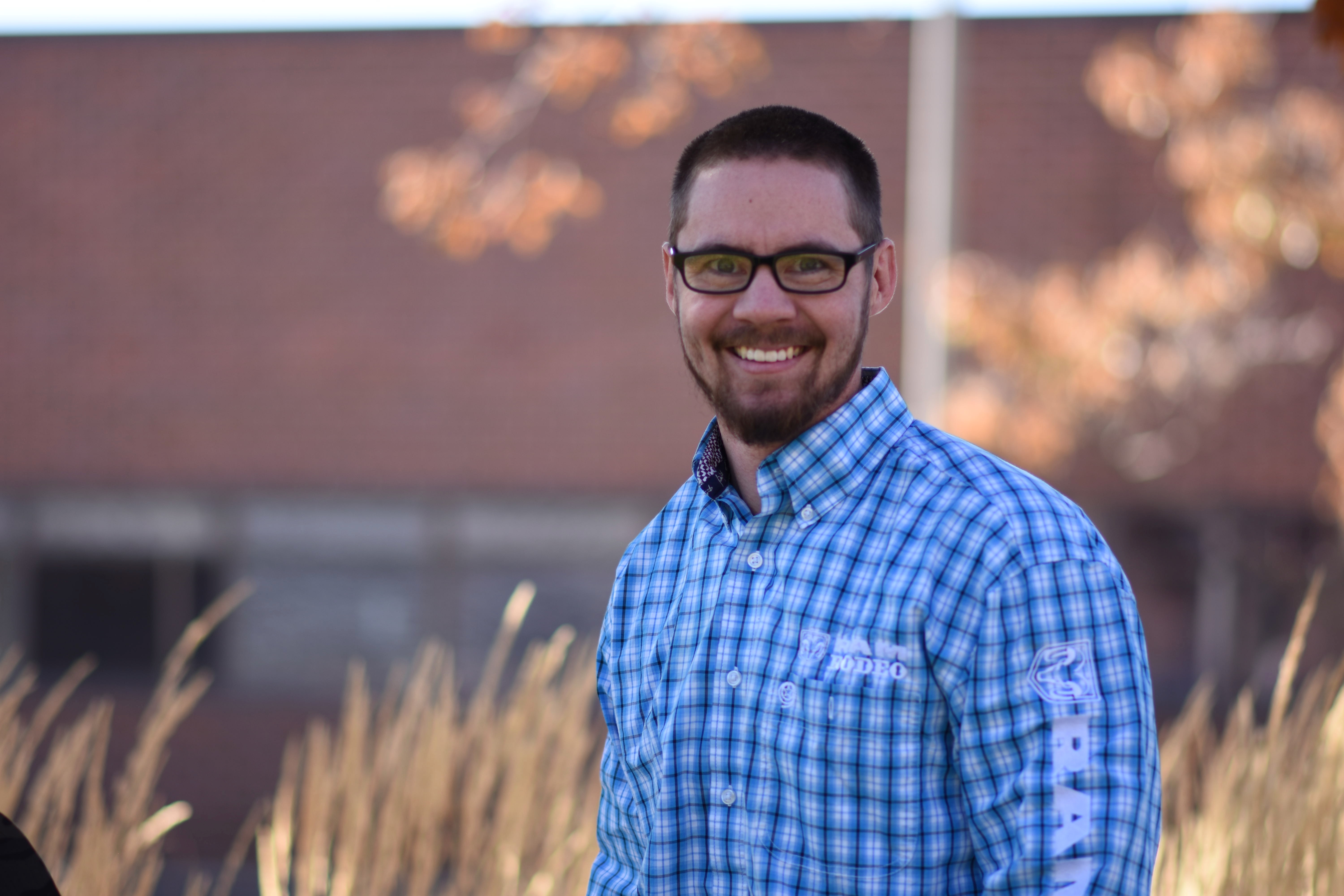 Young white man standing smiling in a blue and white checked shirt.