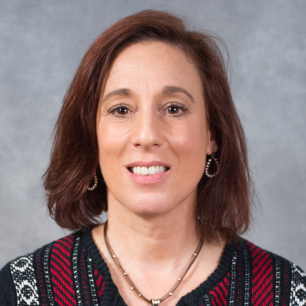 Person with medium length auburn hair wearing patterned top, necklace and hoop earrings