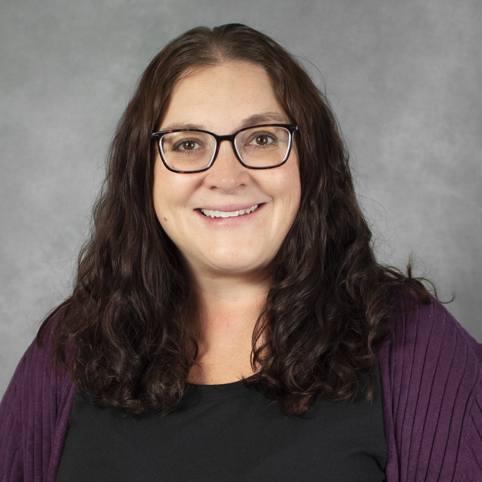 Smiling person with shoulder length dark brown hair wearing glasses, black top and purple sweater