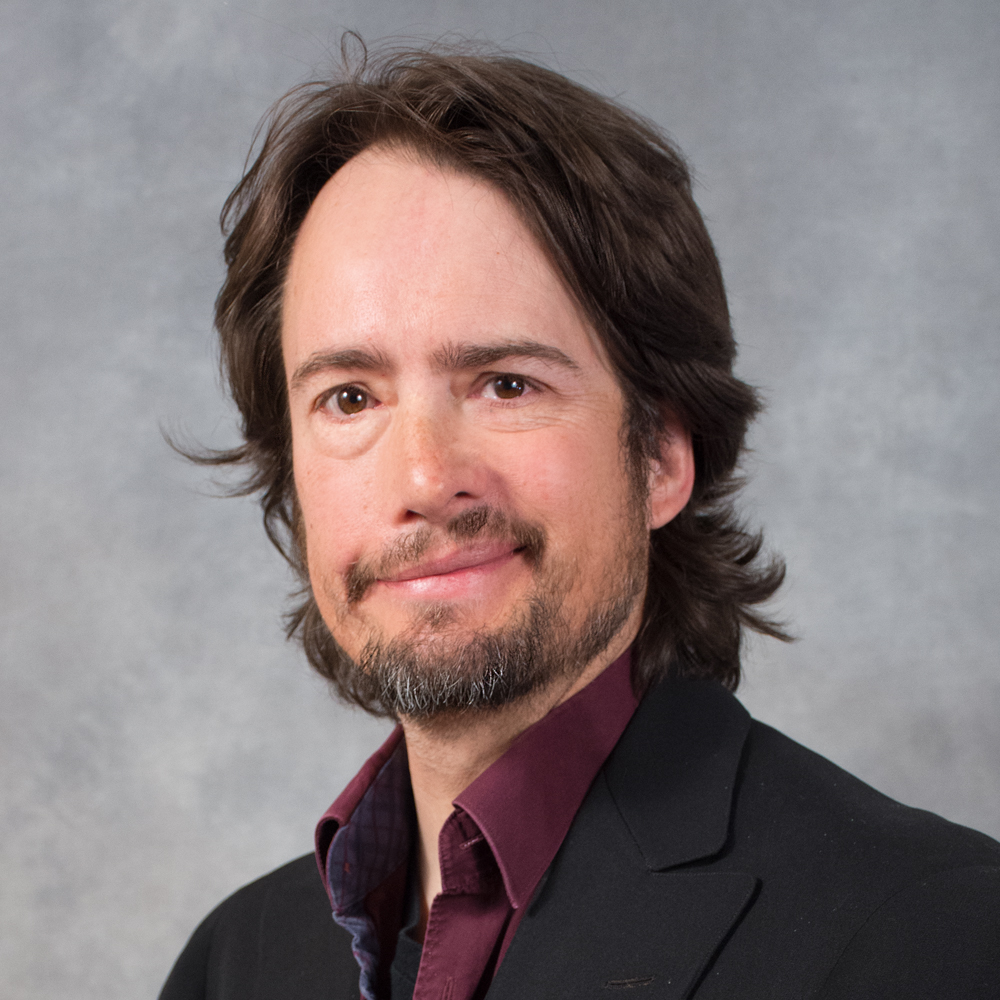 Man with dark brown hair, beard and mustache wearing a dark jacket and burgundy shirt