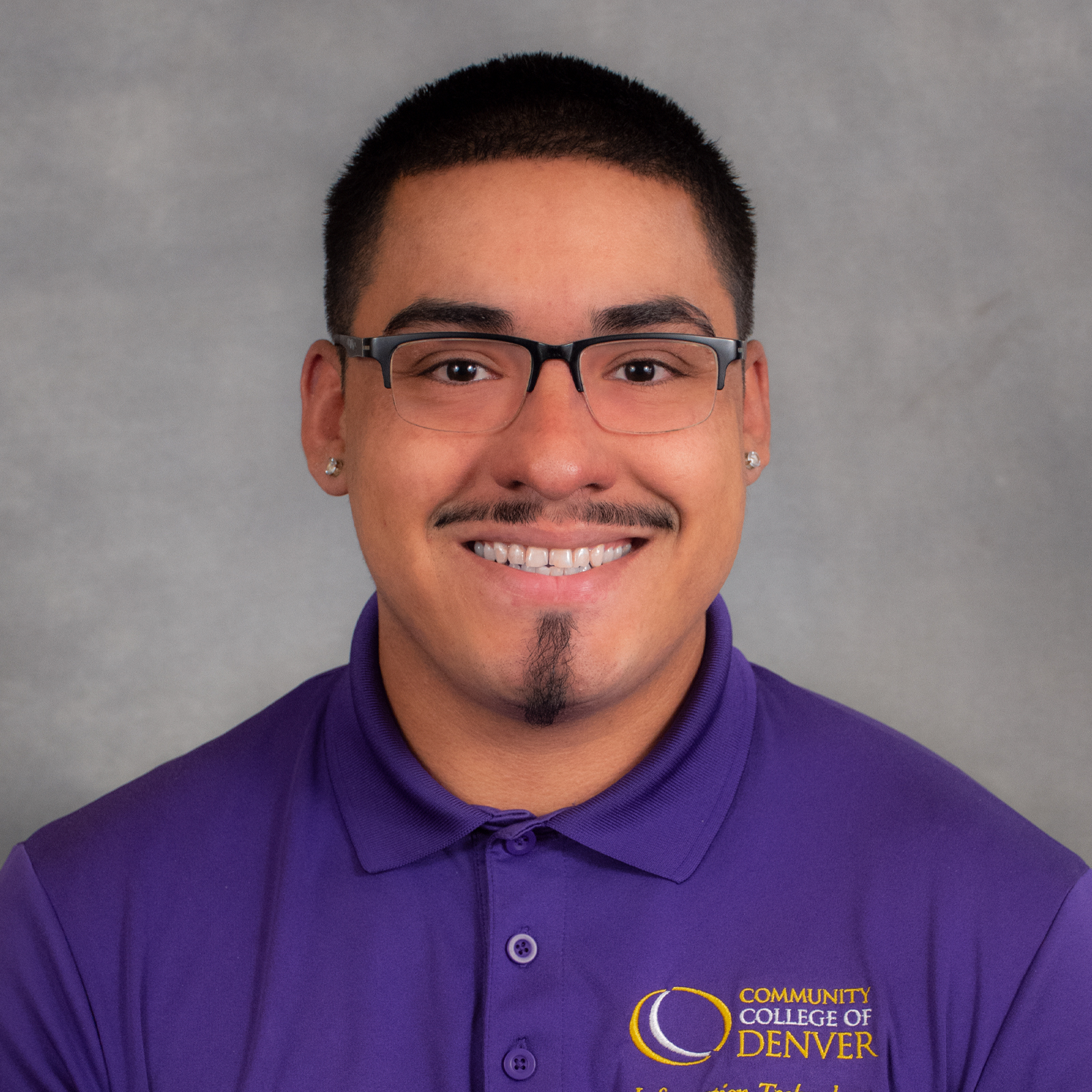 Smiling man with short dark hair, mustasche and chin patch wearing a purple shirt