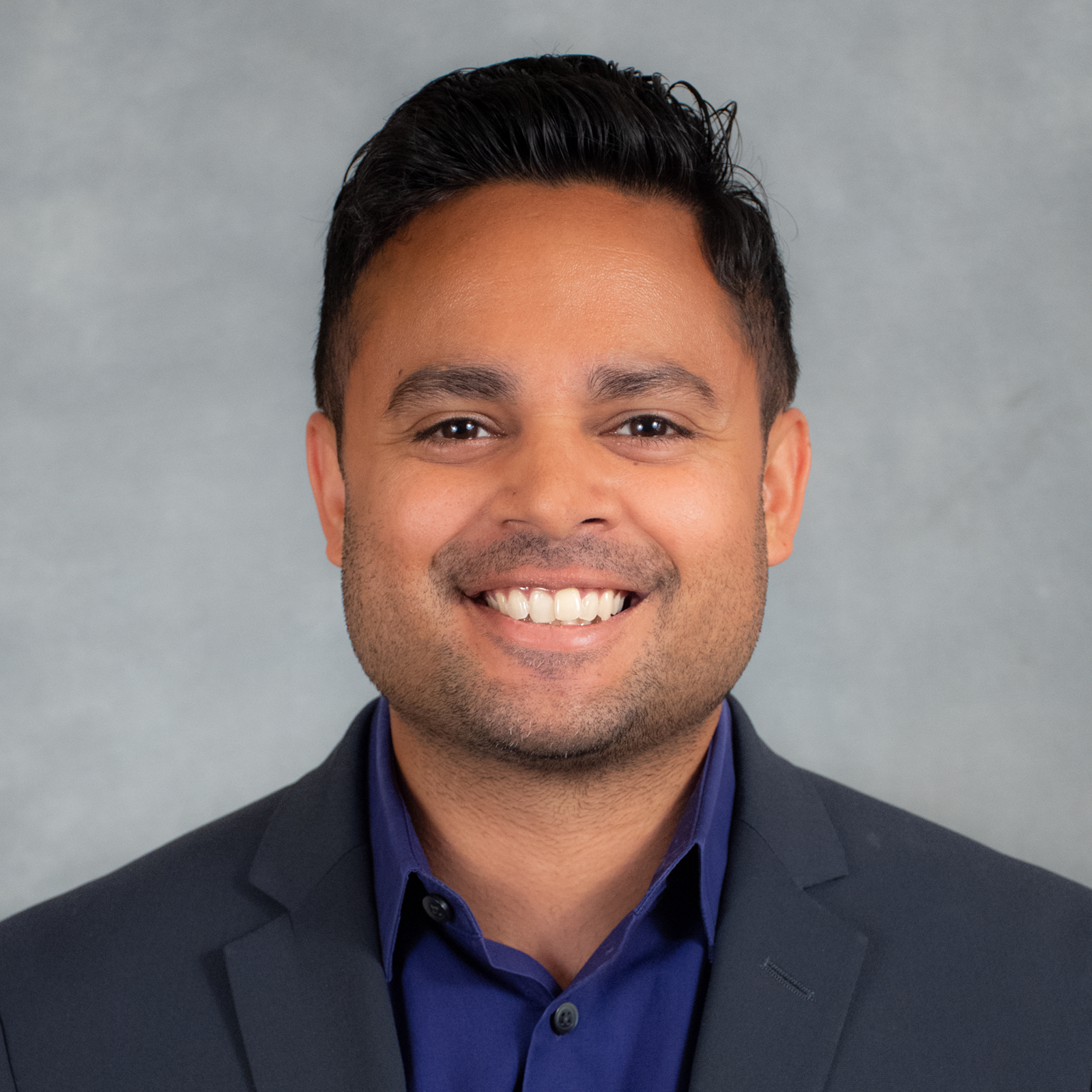 Smiling person with short dark hair wearing dark blue shirt with dark gray suit coat