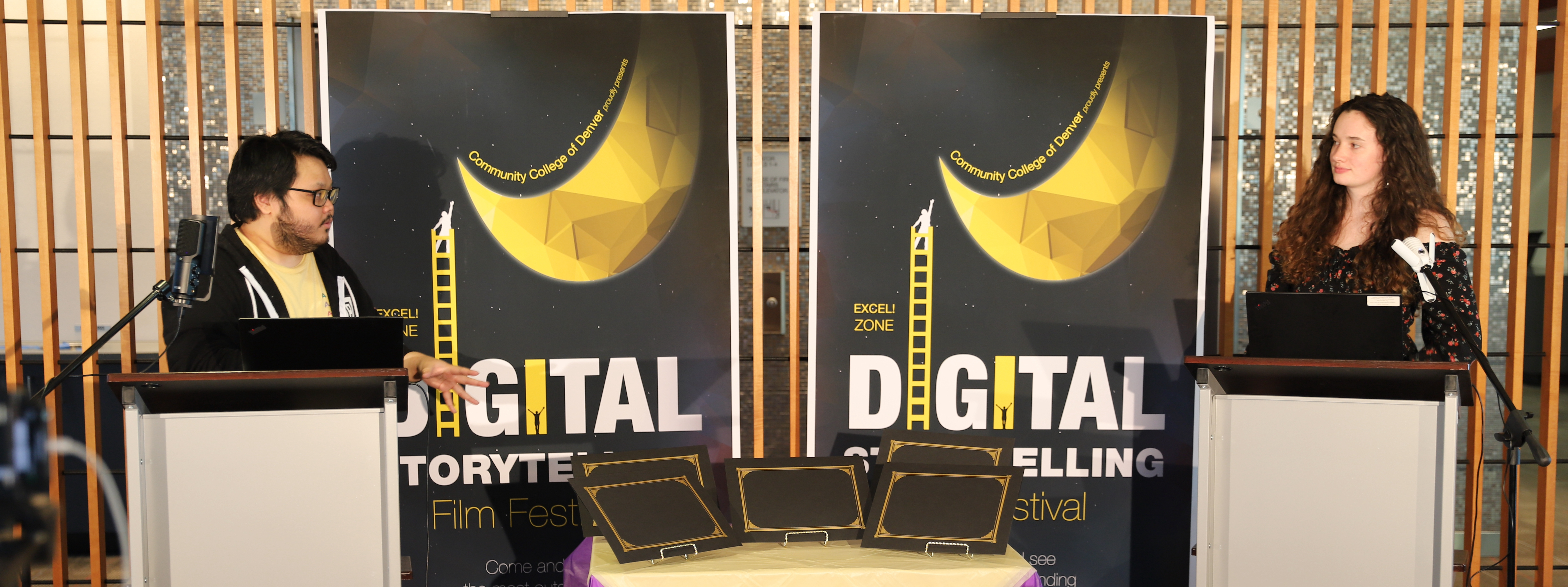 Digital Zone coaches Henry and Claire commentate on the 2021 digital stories Two speakers at two podiums illuminated by hot lights with signs behind them reading "Digital Storytelling Film Festival"