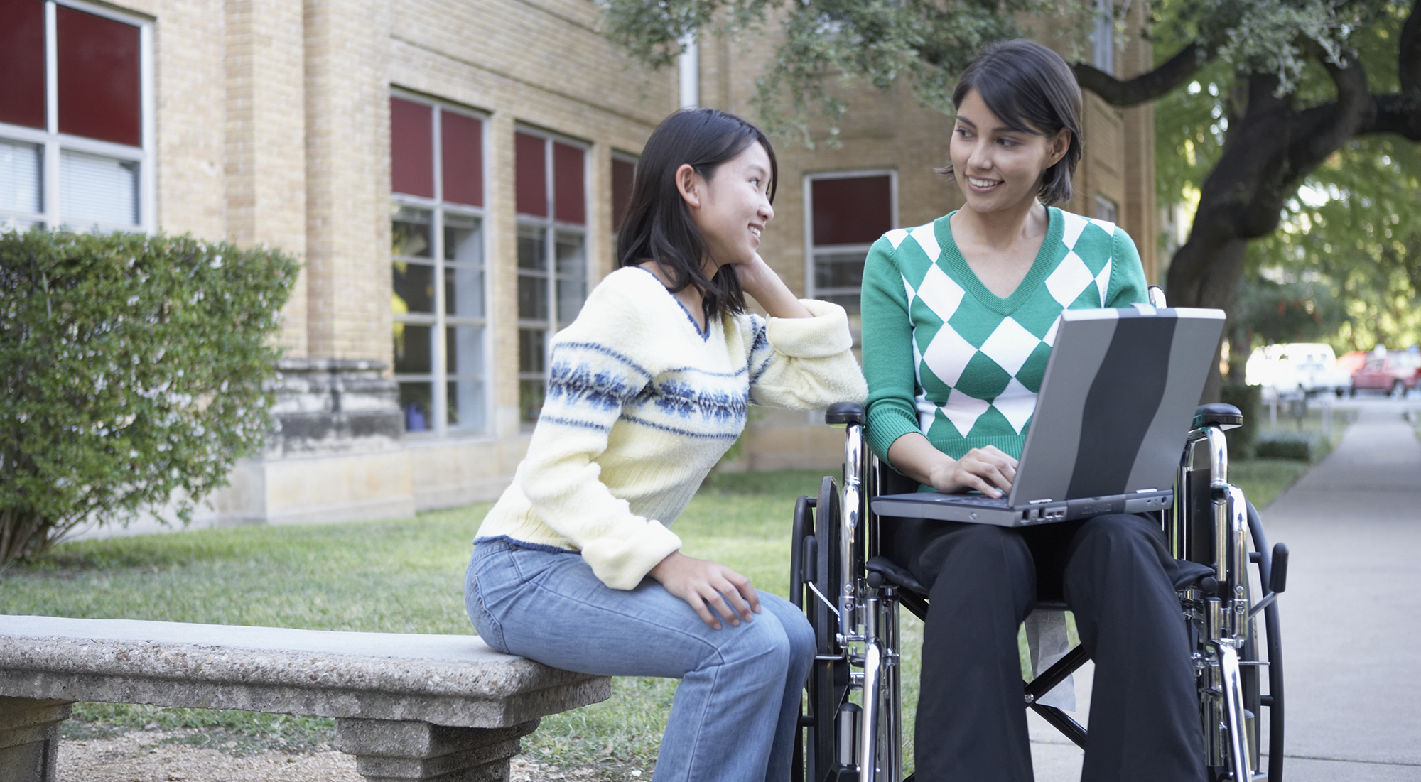 One female student sitting on a concrete bench studying with another female student in a wheelchair holding a laptop on her lap