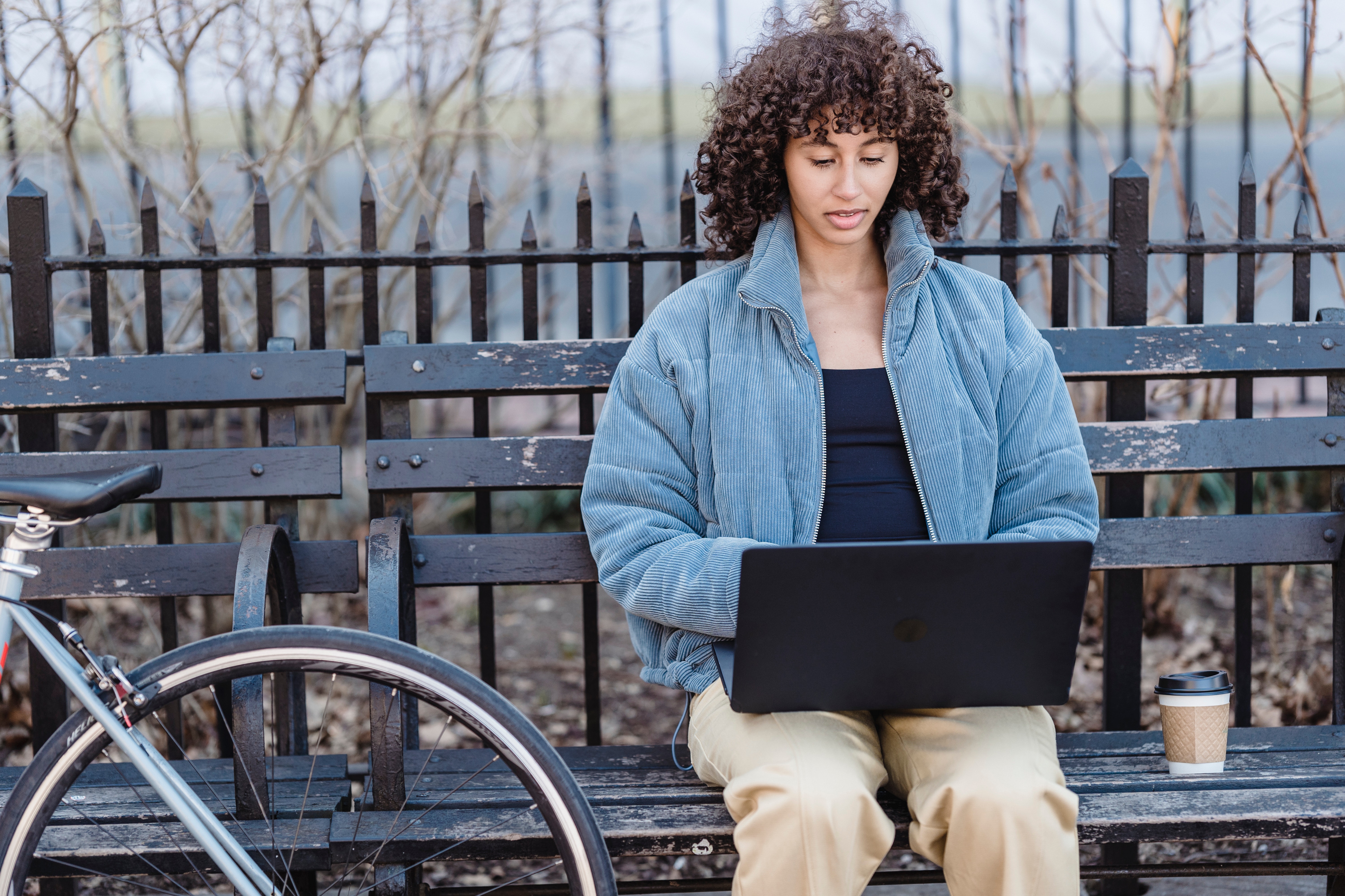 Woman sitting on park bench working on a laptop next to a coffee and a bicycle