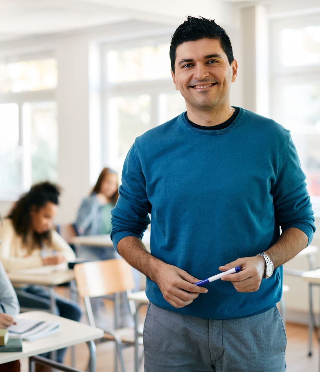 male teacher with black hair a blue shirt holding a pen and smiling in front of a classroom of students studying at their desks