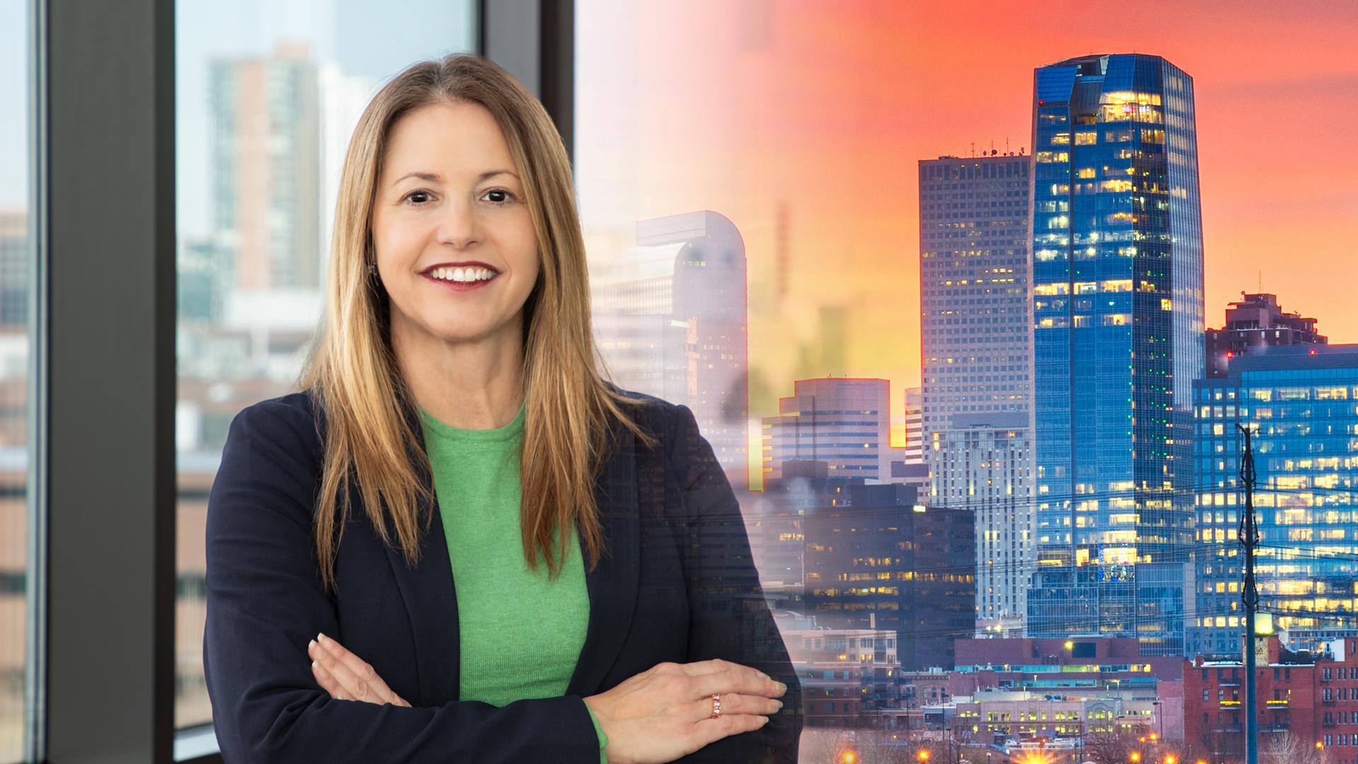 Woman with brown hair wearing a green shirt and blue jacket. Denver skyline in the background.