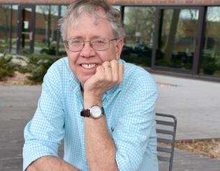 a picutre of a man in a light blue shirt leaning on his hand smiling