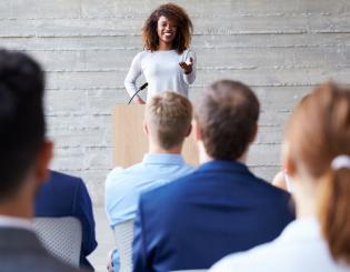 Smiling woman standing at podium giving speech to a seated audience.