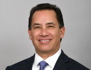 jim chavez smiling and wearing a blue blazer, white collared dress shirt, and light blue tie