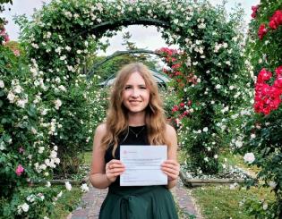 young white woman in a green dress holding a certificate in a garden surrounded by flowers
