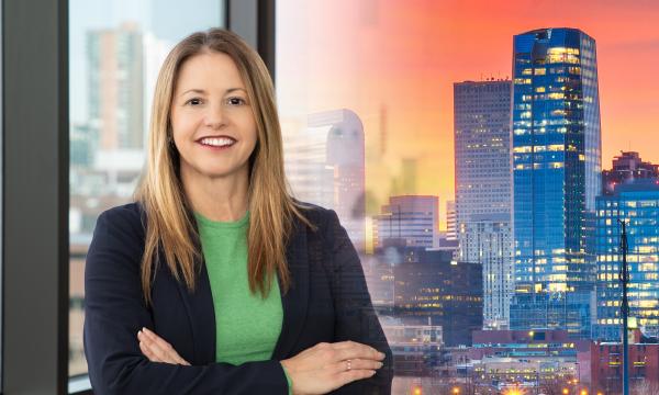 Woman with brown hair wearing a green shirt and blue jacket. Denver skyline in the background.
