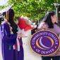 Student wearing purple commencement regalia and holding flowers. A woman wearing a purple shirt stands next to the student. 