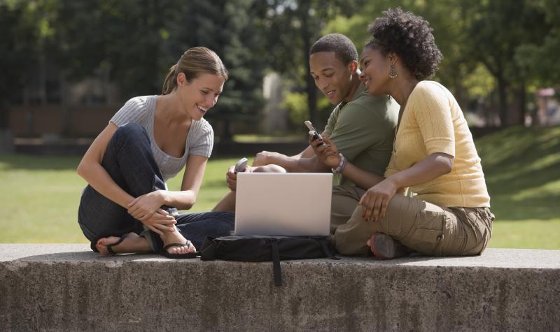 Two female students and one male student sitting outdoors looking at a laptop and phone smiling.