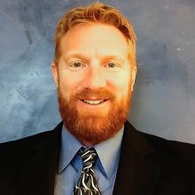 Man with red hair and beard in dark suit, blue shirt and black print tie 