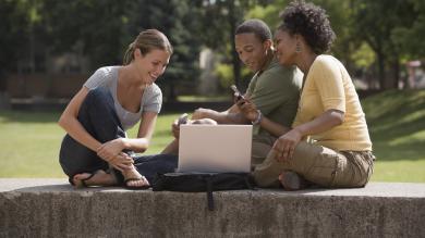Two female students and one male student sitting outdoors looking at a laptop and phone smiling.