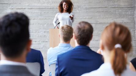 Smiling woman standing at podium giving speech to a seated audience.