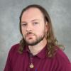 Serious man with shoulder length brown hair, beard and mustache wearing a burgundy shirt