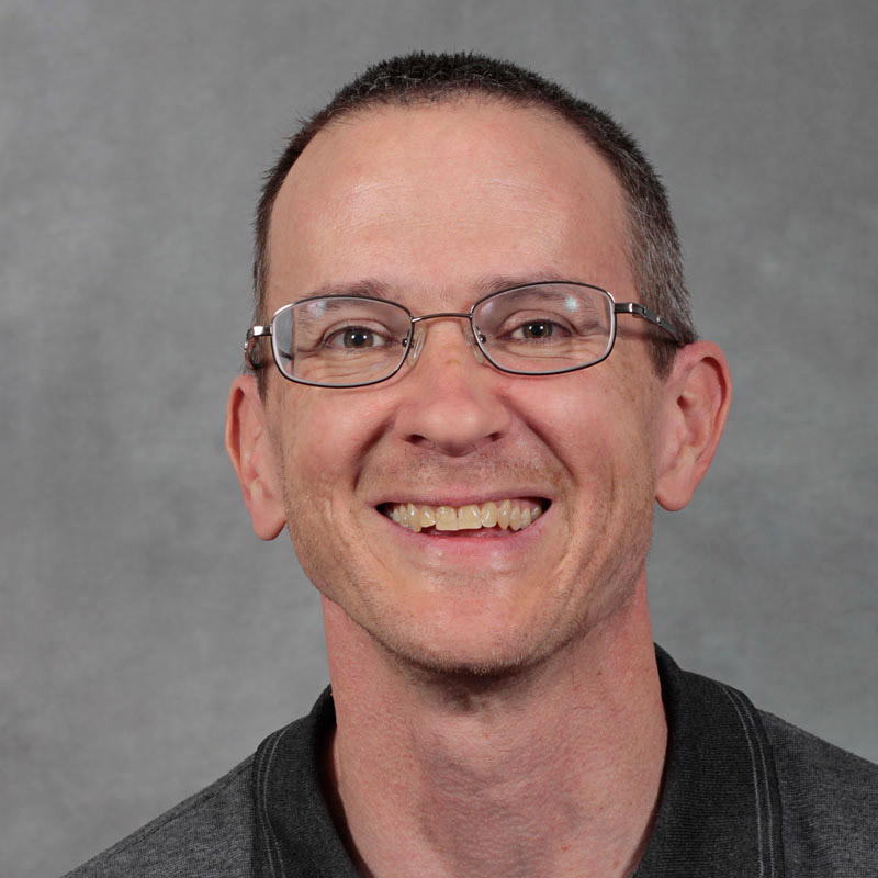 Man with closed cropped dark brown hair, wearing glasses and gray shirt